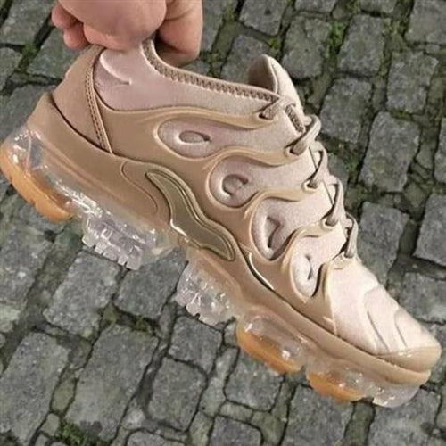 A stylish beige athletic sneaker with a unique, wave-patterned design on the upper, featuring a thick, transparent air-cushioned sole and light brown rubber grip pads. The shoe is being held above a cobblestone pavement.