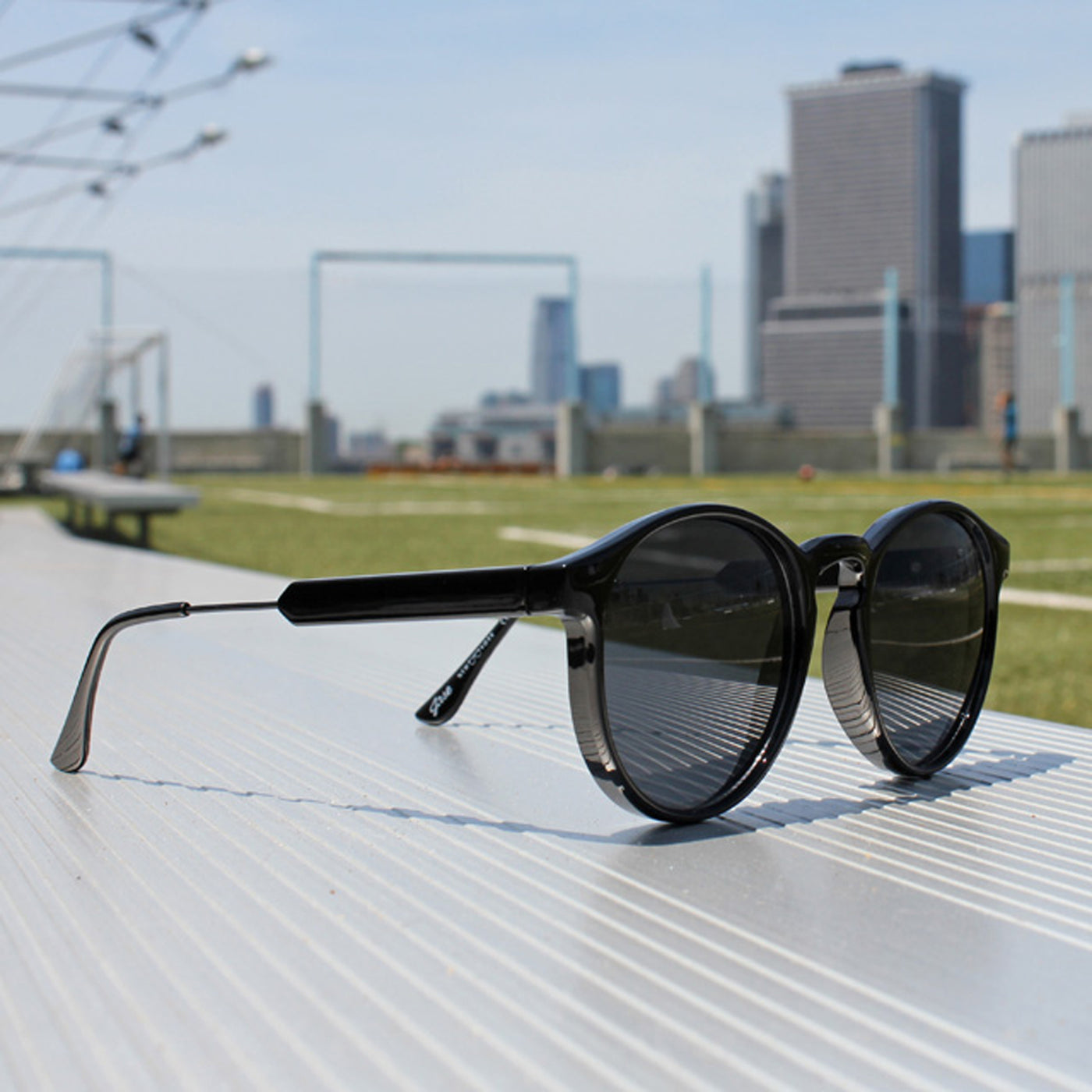 Jase New York Connor Sunglasses in Triple Black displayed outdoors with a city skyline background.