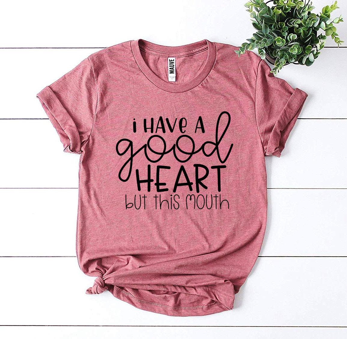 I Have a Good Heart But This Mouth T-shirt displayed on a flat surface with greenery.