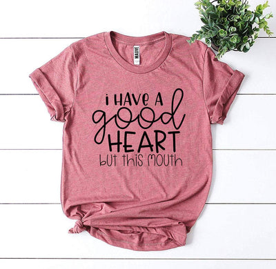I Have a Good Heart But This Mouth T-shirt displayed on a flat surface with greenery.